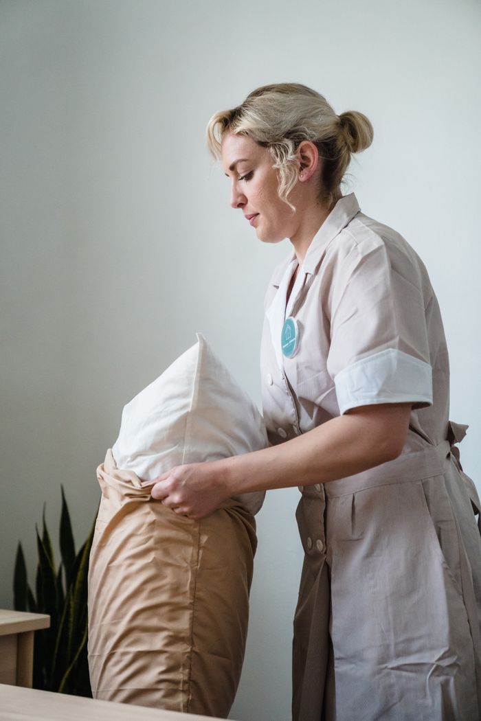 Hotel maid in uniform straightening pillows in a carefully styled guest room.
